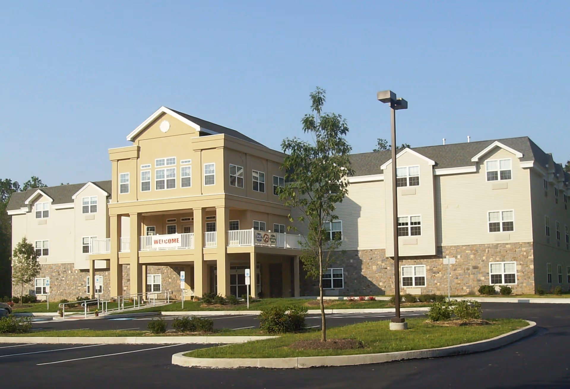 Exterior view of a three-story senior living facility building with beige and stone facade, multiple windows, a covered entrance with columns, a 'WELCOME' banner, a parking lot, and a small tree in front under a clear blue sky.
