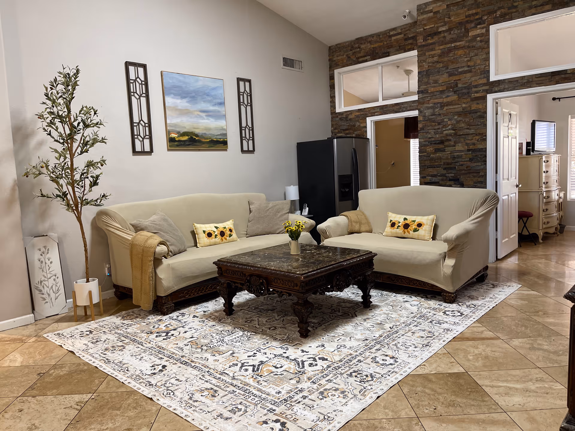 Two beige sofas facing a carved wooden coffee table on a patterned rug in a living room with wall art and a tall potted plant.