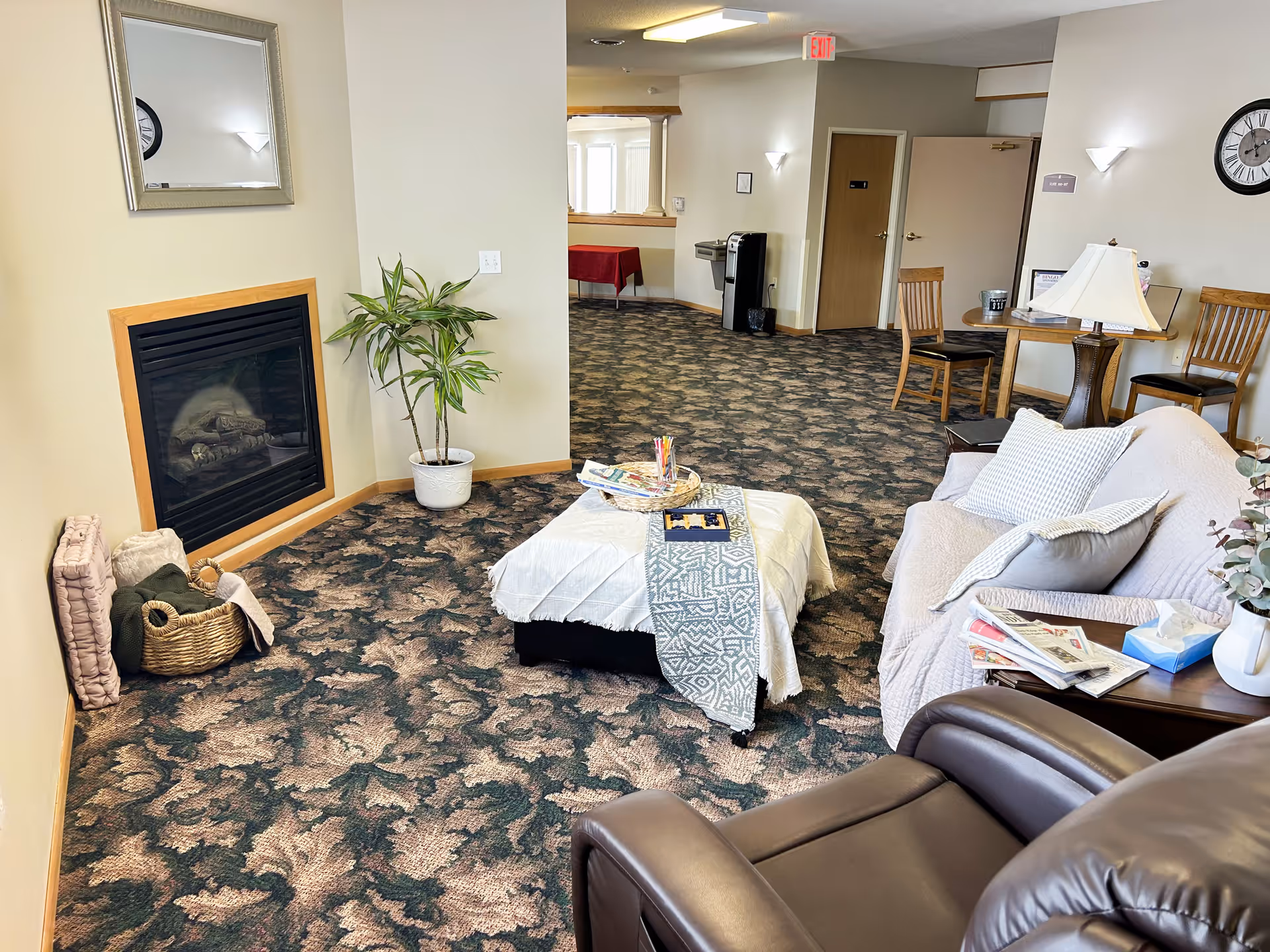 A cozy communal living area with sofas, an ottoman, a fireplace, chairs and tables on patterned carpet.