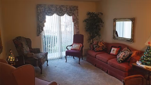 A cozy living room with a red sofa adorned with floral and holiday-themed pillows, two armchairs, a small decorated Christmas tree on a side table, a large window with vertical blinds and floral valance, a potted plant in the corner, and a framed mirror on the wall.