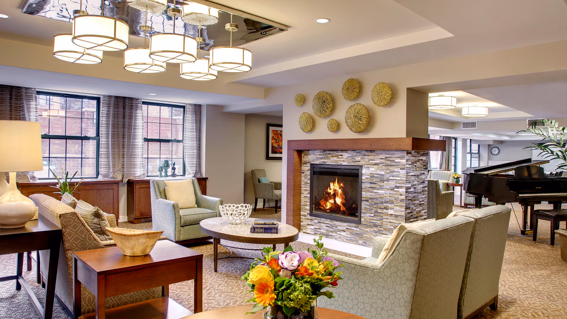 Comfortable senior living common room with sofas and armchairs arranged around a lit fireplace, decorative wall art, a coffee table with flowers, and a grand piano in the background.