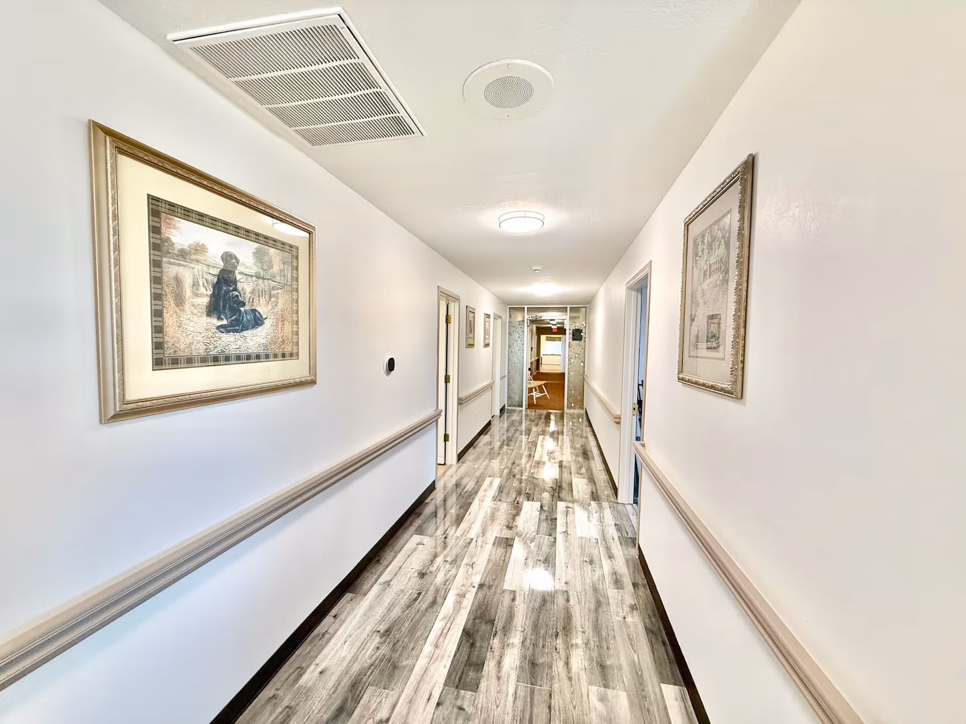Bright polished hallway in a senior living facility with framed artwork on the walls and wood-look flooring.