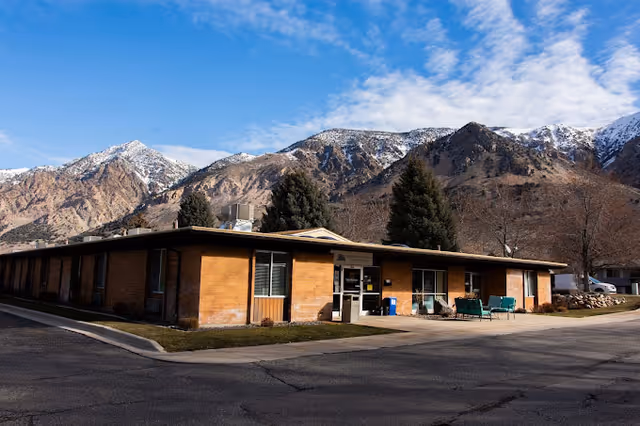 Single-story brick building with large windows and a flat roof, situated in front of a mountainous landscape with snow-capped peaks under a partly cloudy blue sky. There are a few evergreen trees near the building and outdoor seating with green chairs on the right side of the entrance.