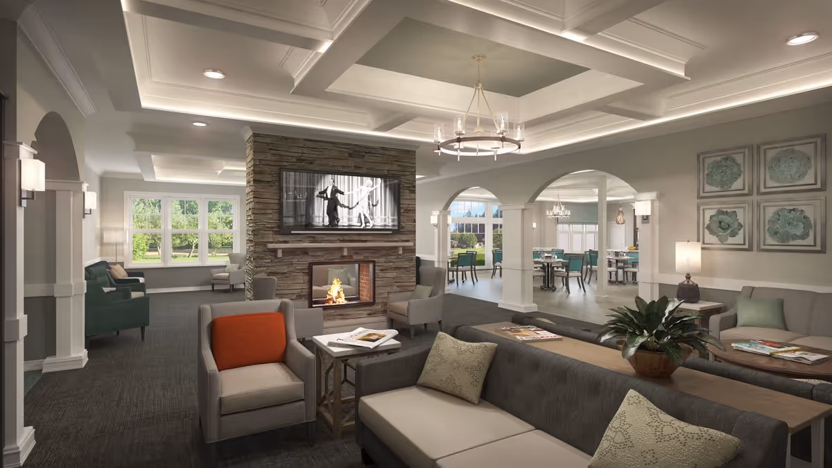 A spacious and well-lit senior living facility common area featuring comfortable seating with armchairs and sofas arranged around a stone fireplace with a mounted TV above it. The room has large windows letting in natural light, decorative ceiling with recessed lighting, and archways leading to a dining area with tables and chairs. The walls are adorned with framed artwork and there are plants and lamps adding to the cozy ambiance.