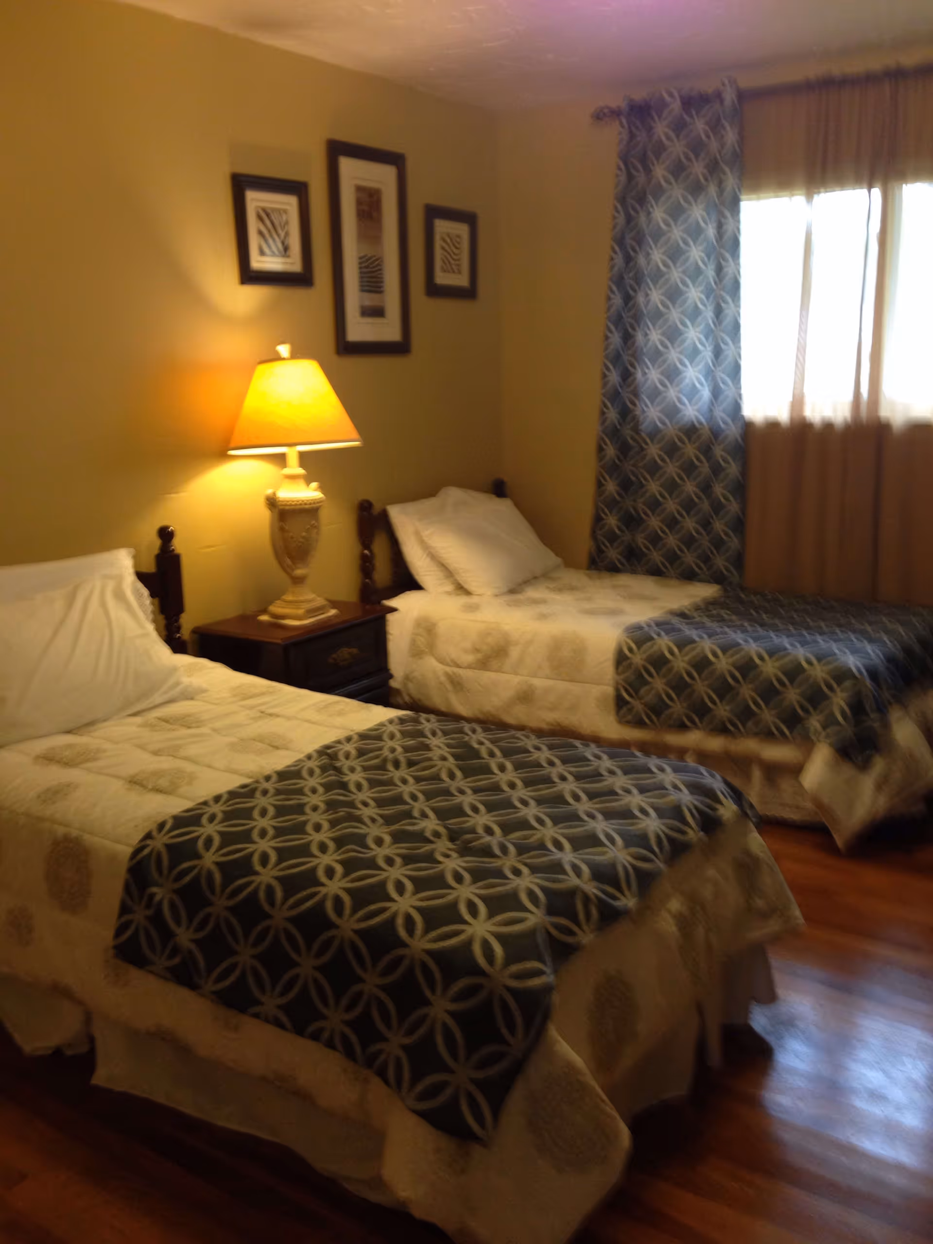 A bedroom with two twin beds, each with white and patterned bedding. Between the beds is a wooden nightstand with a decorative lamp. The walls are painted beige and have three framed pictures. A window with patterned and sheer curtains lets in natural light.