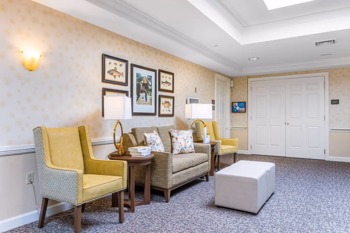 A well-lit sitting area in a senior living facility with two yellow armchairs, a beige sofa with floral cushions, two side tables with lamps, framed artwork on the wall, and a white ottoman on a carpeted floor.