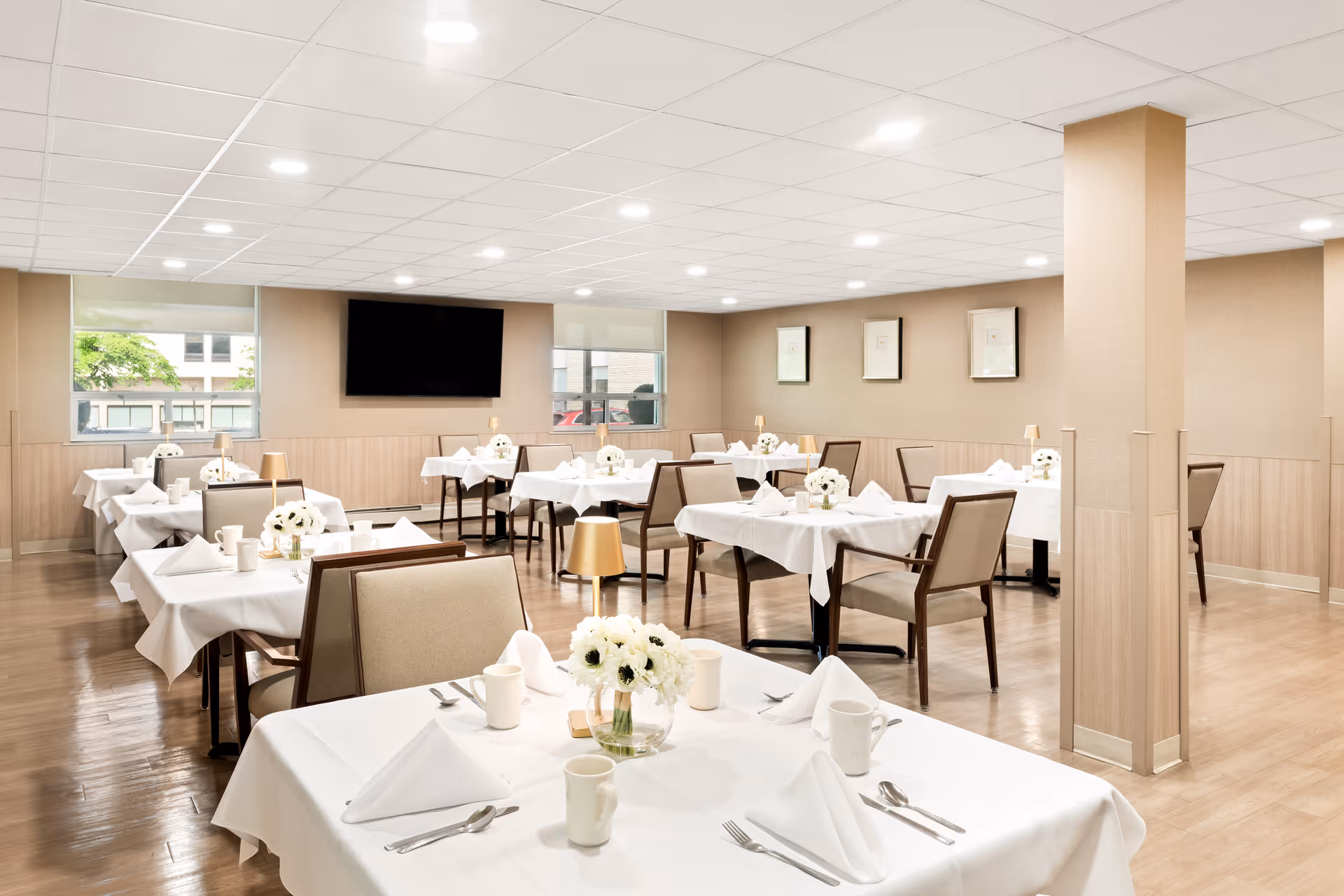 Bright, neatly arranged dining room with white-tablecloth tables, beige chairs, floral centerpieces and a wall-mounted TV.