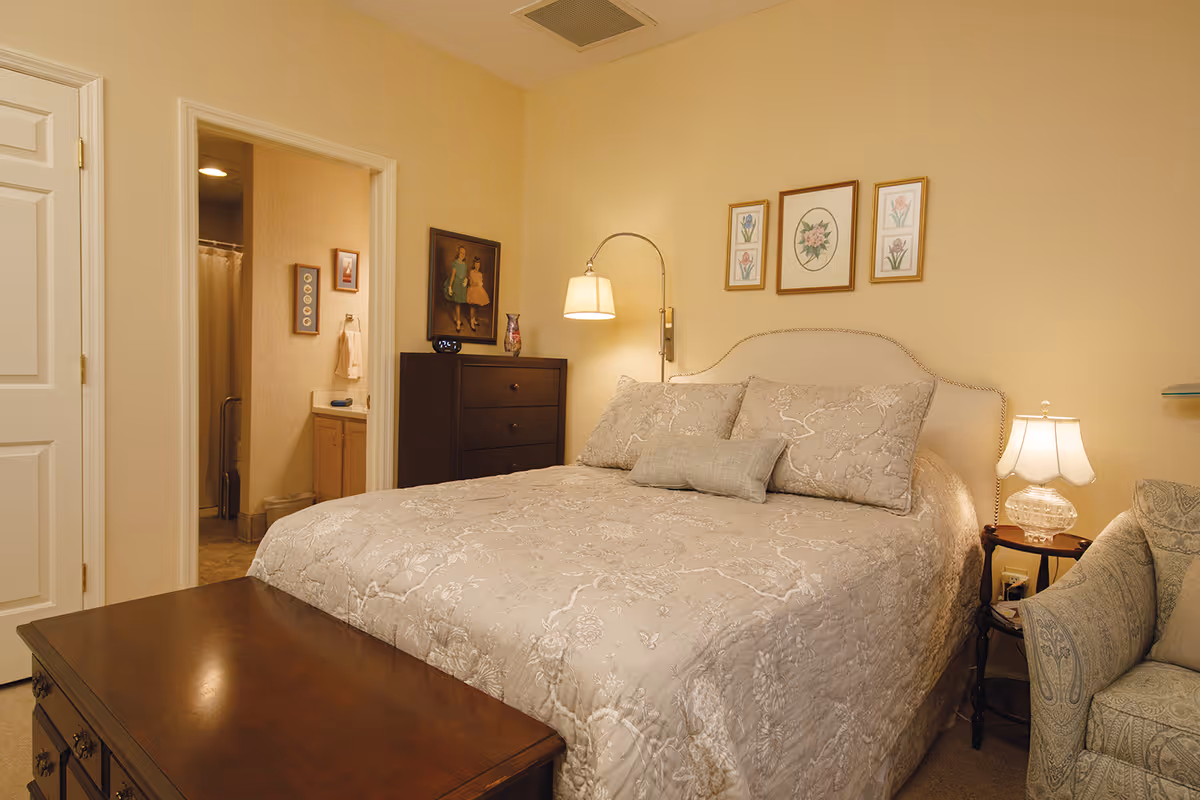 A cozy bedroom with a large bed covered in a beige floral quilt and matching pillows. There is a wooden chest at the foot of the bed, a dark wooden dresser with a decorative vase and a framed picture above it, and a wall-mounted lamp beside the bed. On the right side, there is a small side table with a glass lamp and a patterned armchair. An open doorway reveals a bathroom with a shower curtain and a sink.