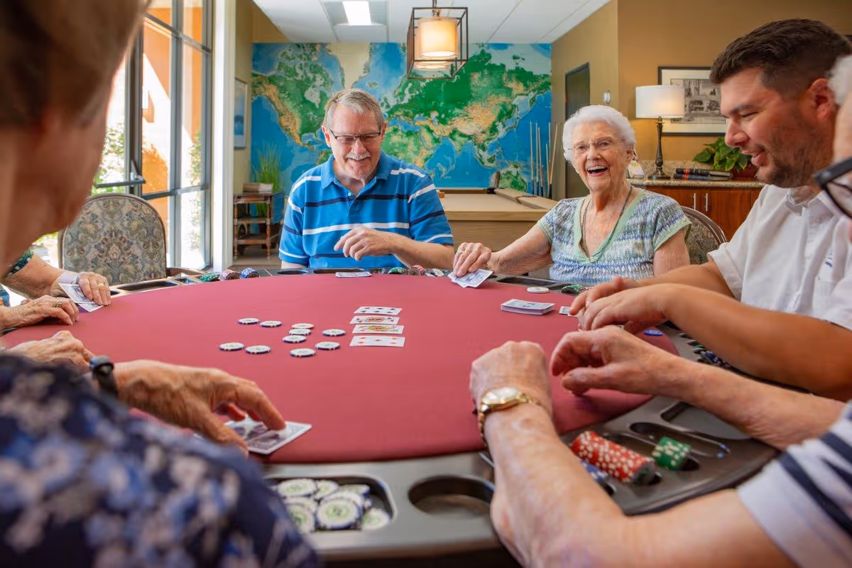 A group of elderly people and a younger man sitting around a red poker table playing cards and poker chips. They are smiling and enjoying the game in a well-lit room with a large world map on the wall and a pool table in the background.