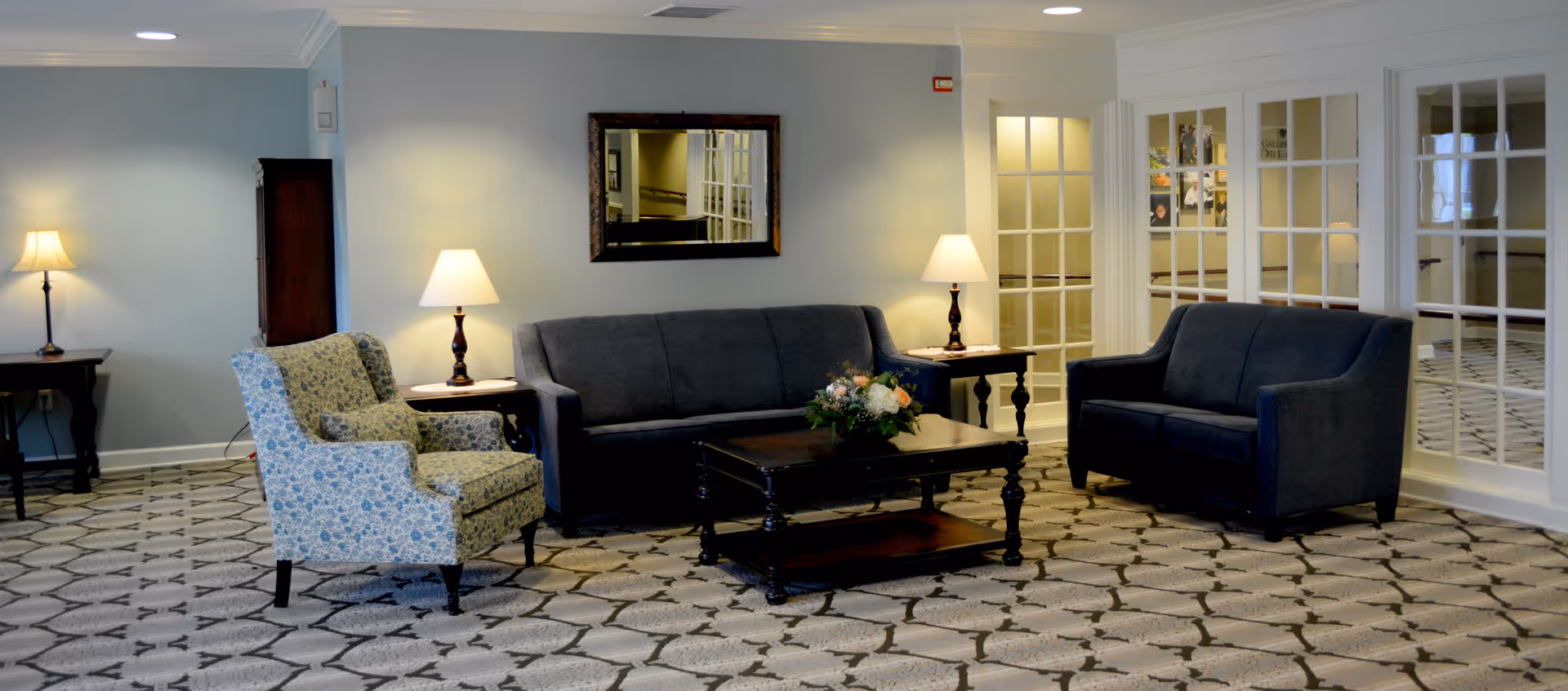A well-lit living room area with a patterned carpet, two dark blue sofas, a floral upholstered armchair, a wooden coffee table with a flower arrangement, two side tables each with a lamp, and a large mirror on the wall. There are glass-paneled doors on the right side of the room.