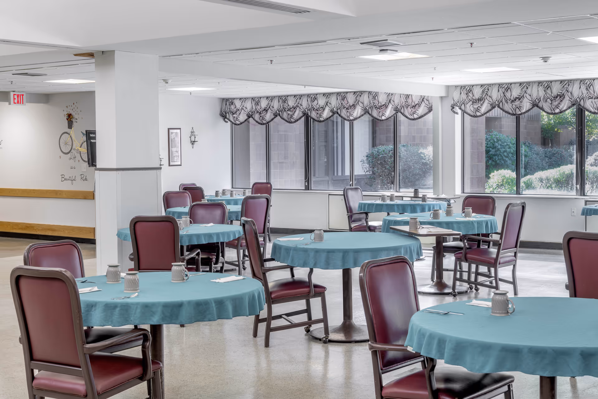 A dining room with multiple round tables covered with teal tablecloths, each surrounded by maroon cushioned chairs. The tables have mugs and napkins placed on them. Large windows with patterned valances allow natural light to brighten the room, and some greenery is visible outside.