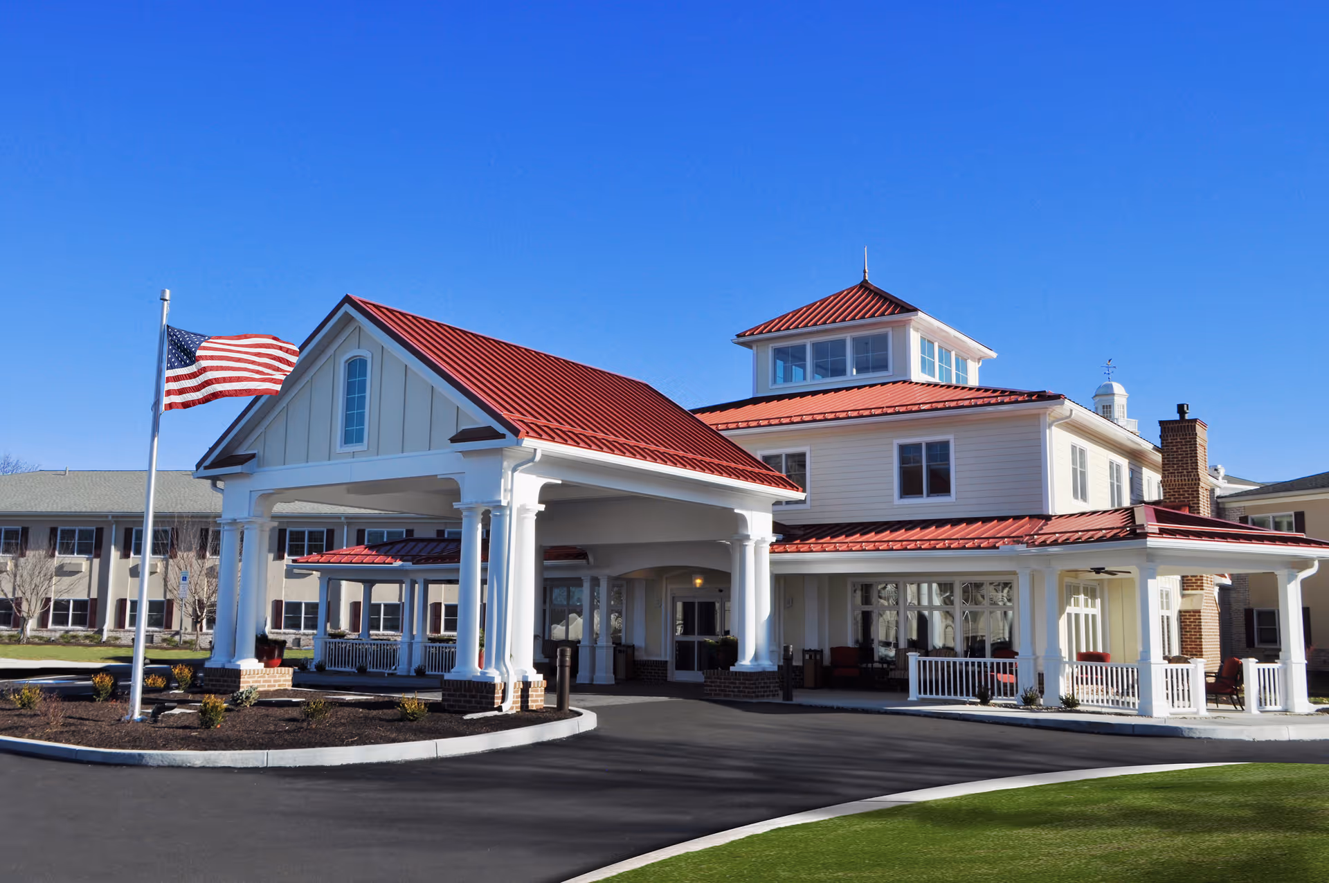 Front entrance of a large retirement community building with a red metal roof, white columns, and an American flag flying out front.