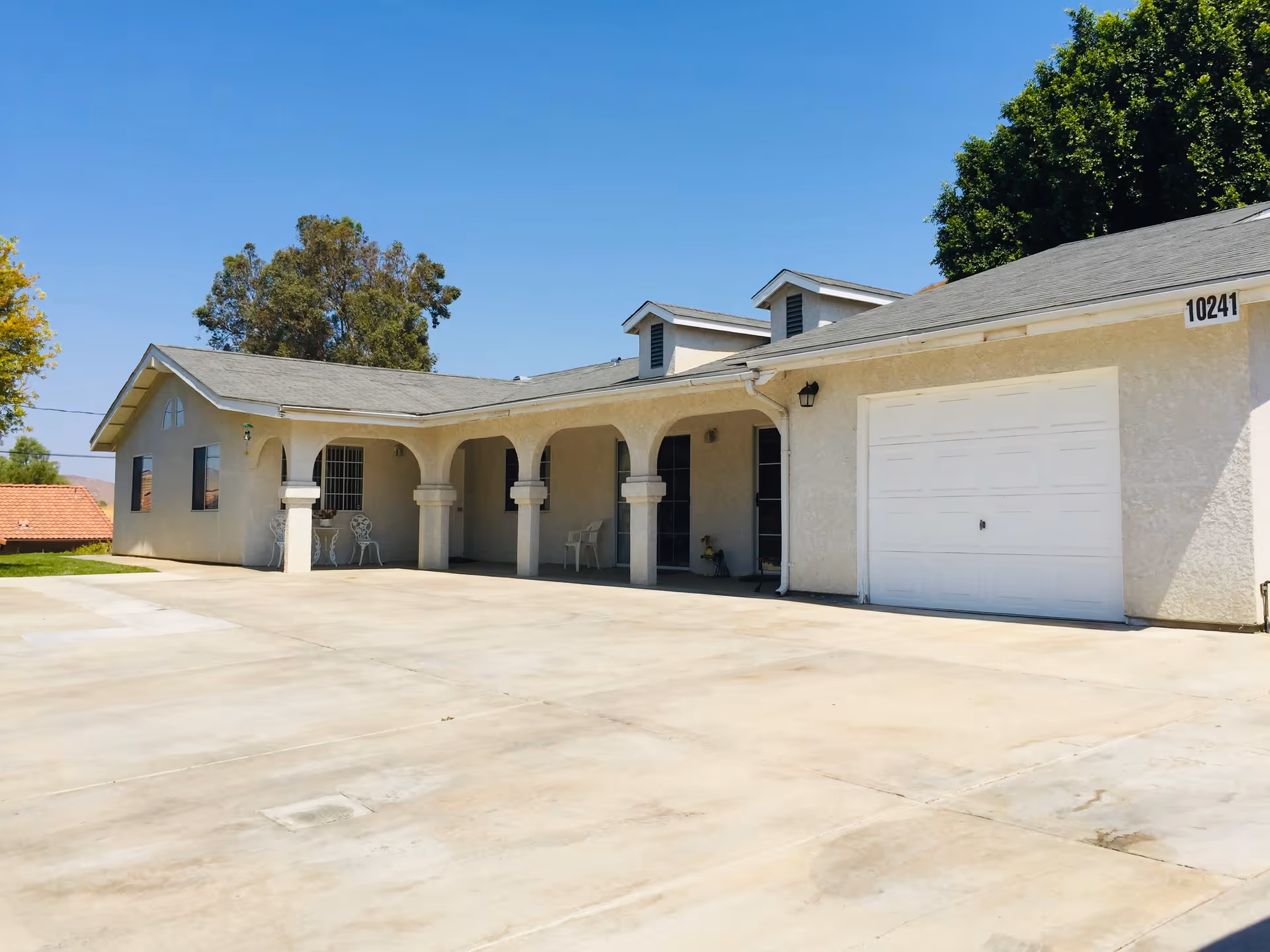 Exterior view of a single-story building with a light-colored facade, arched porch supported by columns, a white garage door, and a concrete driveway under a clear blue sky. Trees and a neighboring roof are visible in the background.
