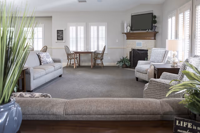 A cozy living room with gray sofas and patterned armchairs arranged around a stone fireplace with a mounted TV above it. There is a wooden table with two chairs near the windows with white shutters, and several green plants add a touch of nature to the room. A lamp on a side table provides soft lighting.