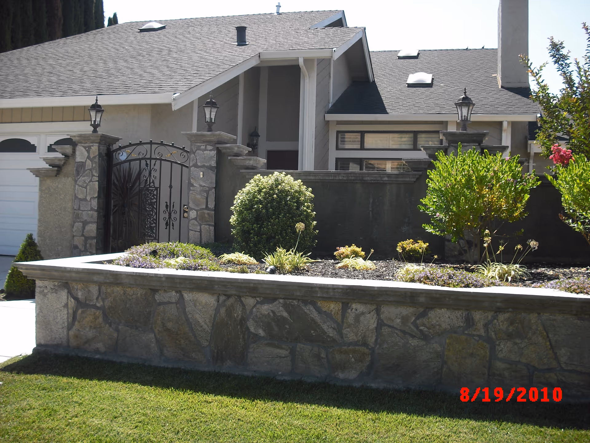 Front exterior view of a single-story house with a stone wall and gate in front, surrounded by a well-maintained garden with shrubs and small plants. The house has a gray roof and beige walls, with a garage door visible on the left side.