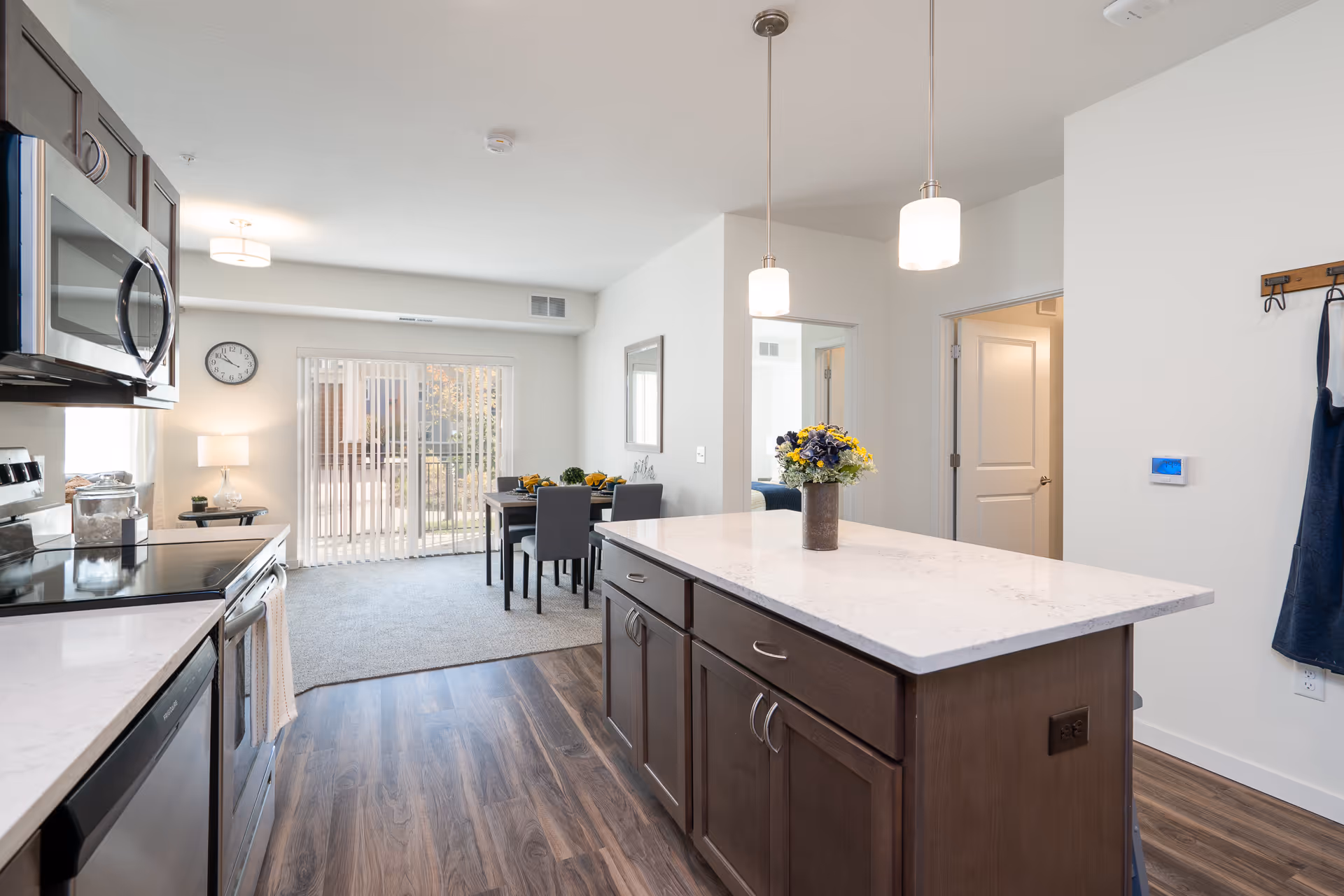 Bright and modern kitchen area with dark wood cabinets and a white marble countertop island. Stainless steel appliances including a microwave and stove are visible on the left. Two pendant lights hang above the island. In the background, there is a dining area with a table and chairs near sliding glass doors with vertical blinds, allowing natural light to enter. The floor transitions from wood in the kitchen to carpet in the dining area. A clock and a lamp are visible on the far wall.