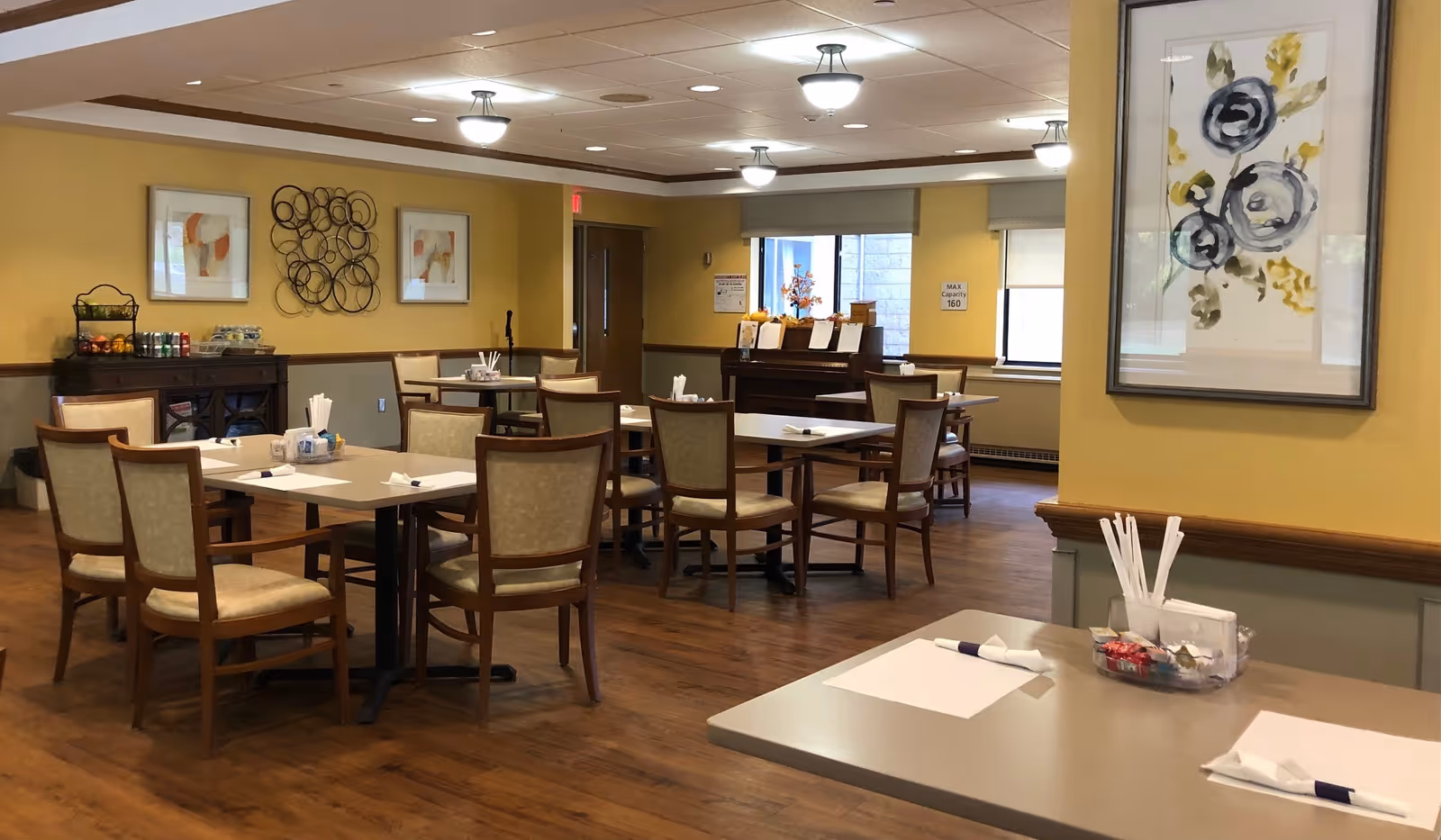 A dining room in a senior living facility with several tables and chairs arranged neatly. The tables are set with napkins and utensils. The room has yellow walls adorned with framed artwork and decorative wall hangings. There is a sideboard with beverages and snacks, and windows letting in natural light.