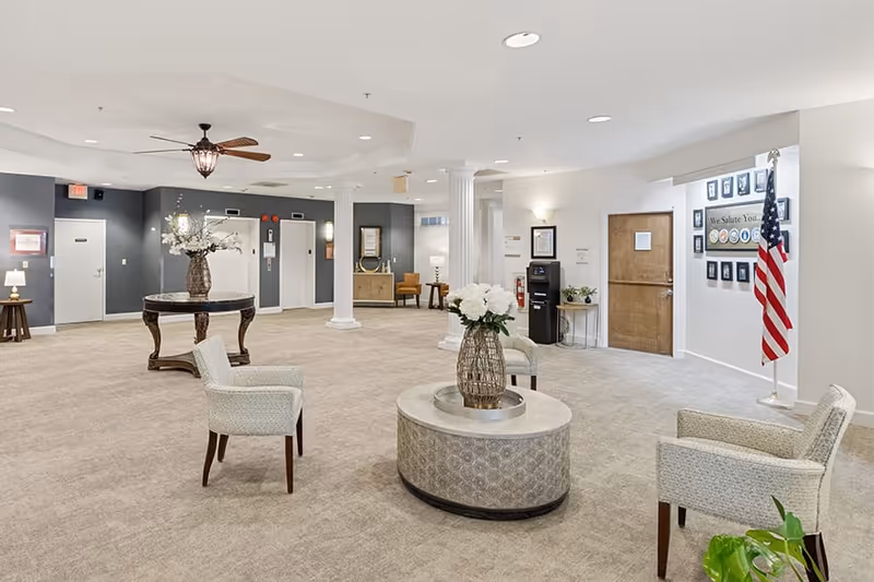 A spacious senior living facility lobby with beige carpet, white walls, and decorative columns. The room features comfortable armchairs arranged around a round table with a vase of white flowers. There is a round wooden table with a large floral arrangement, an American flag, framed pictures on the wall, and a water dispenser. The area is well-lit with ceiling lights and a ceiling fan.