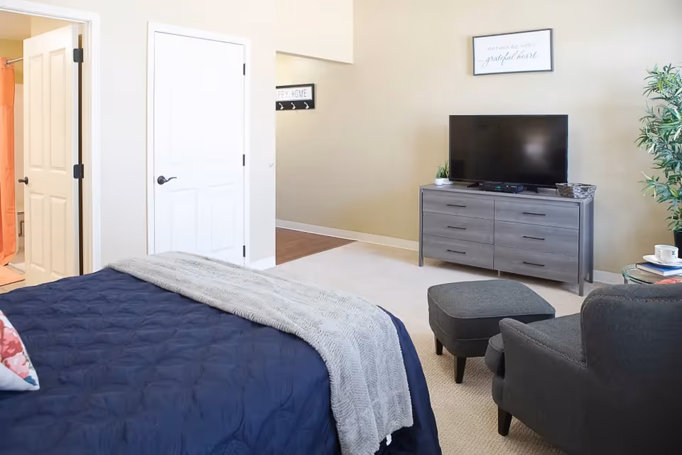 A bedroom with a navy blue bedspread and a light gray blanket. There is a gray armchair with a matching ottoman, a gray dresser with a flat-screen TV on top, and a potted plant next to the dresser. Two white doors are visible, one leading to a bathroom with an orange shower curtain. The walls are beige, and there are framed signs on the wall.