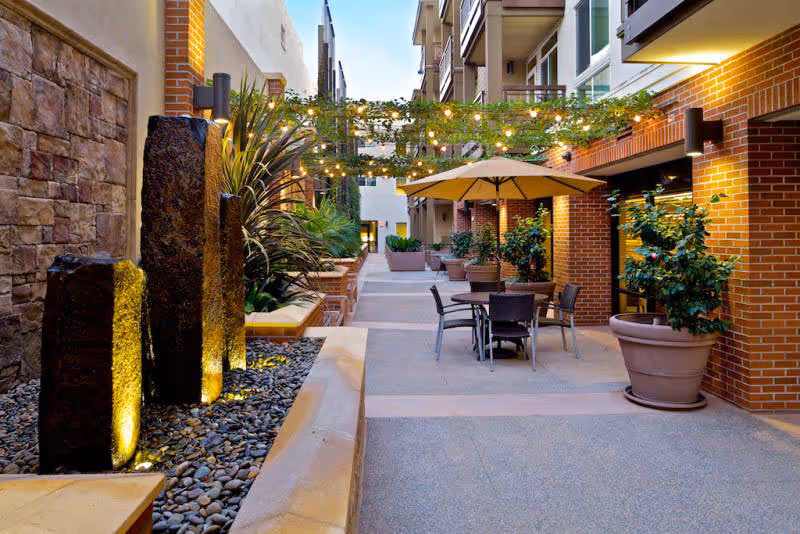 Outdoor courtyard area with string lights overhead, potted plants, a table with four chairs and a large umbrella, and three vertical stone water features illuminated from below.