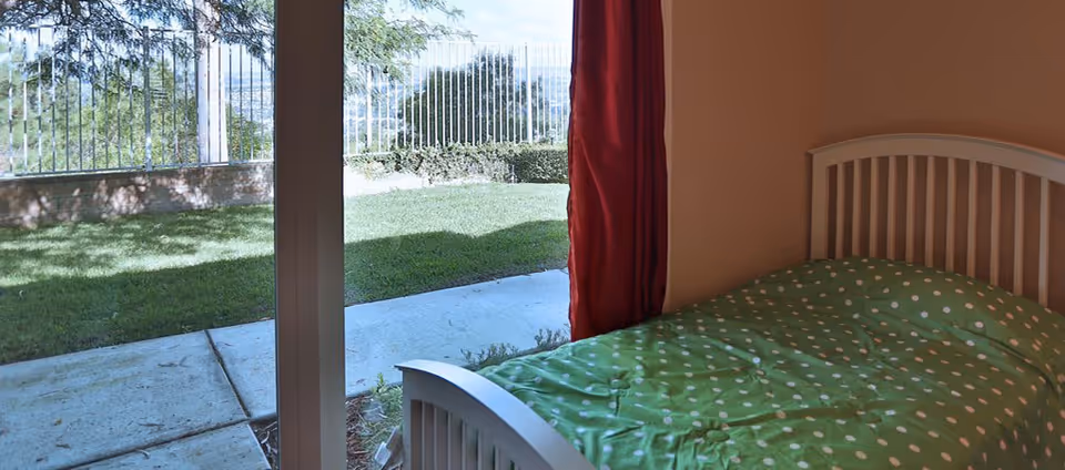 A bedroom with a white wooden bed frame and a green polka dot blanket. The room has a large glass sliding door that opens to a small outdoor area with grass, a concrete path, and a metal fence. A red curtain is partially drawn to the side of the door.