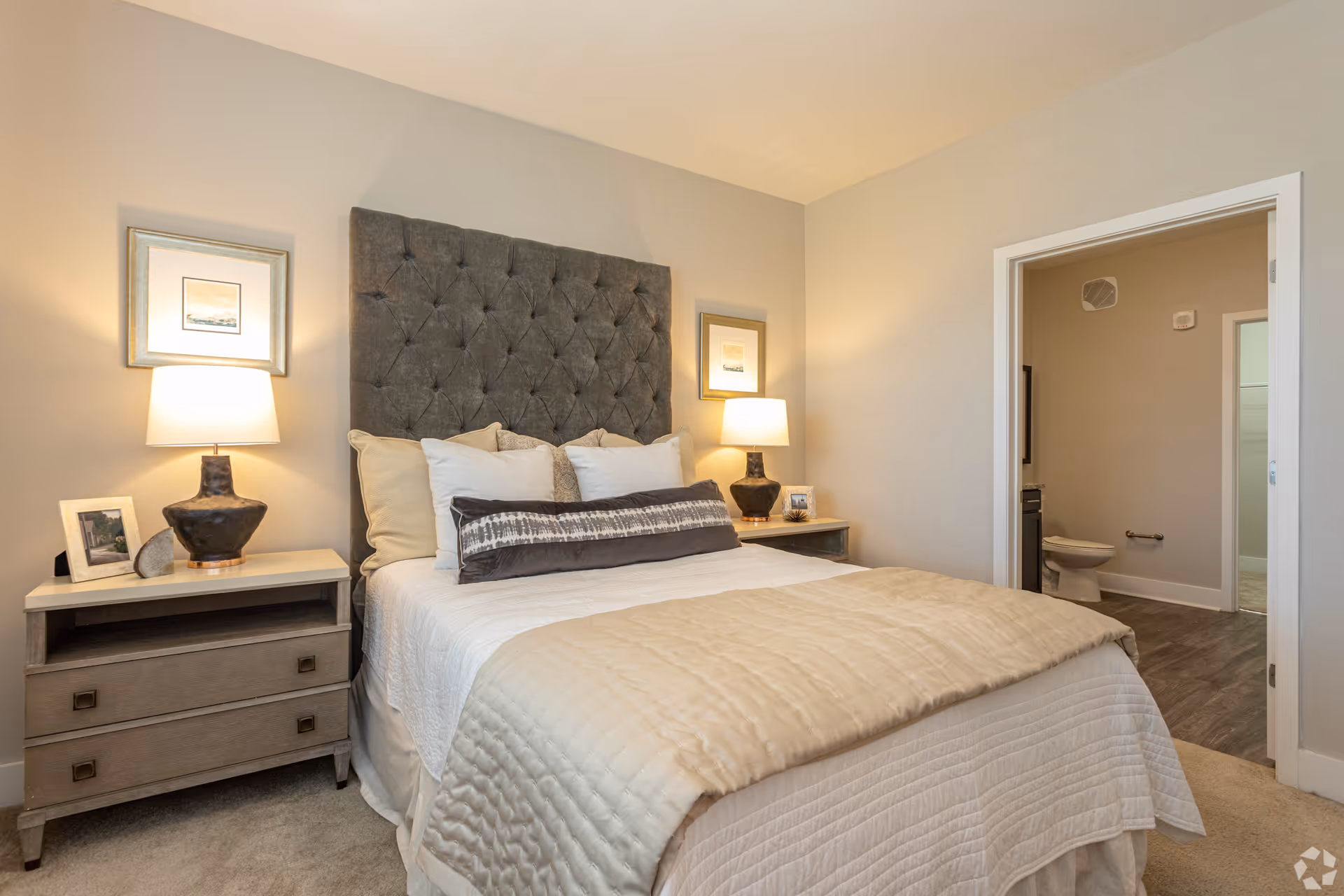 A cozy bedroom with a large tufted gray headboard, a neatly made bed with white and beige bedding, two bedside tables each with a lamp and framed pictures, and an open door leading to a bathroom with a visible toilet.