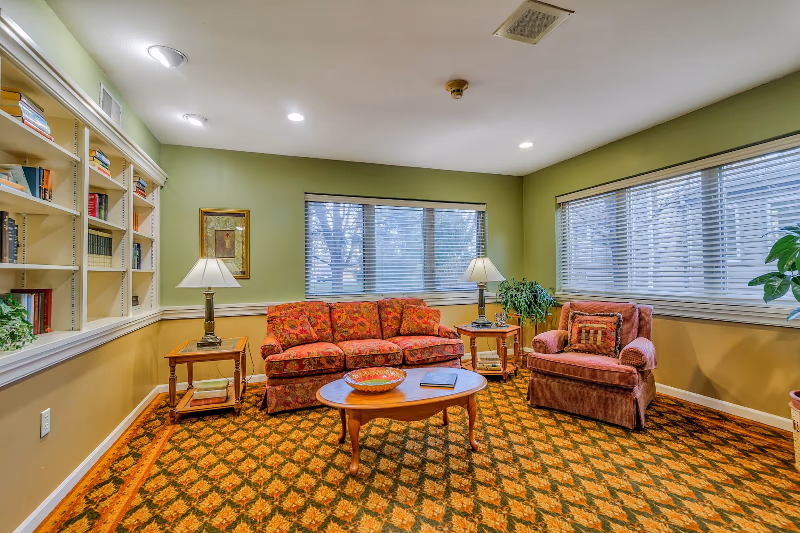 A cozy living room with green walls and large windows with blinds. The room features a floral patterned sofa, a matching armchair with a decorative pillow, two wooden side tables with lamps, a wooden coffee table with a decorative bowl and a book, a built-in bookshelf filled with books, and a patterned carpet with green and orange tones.