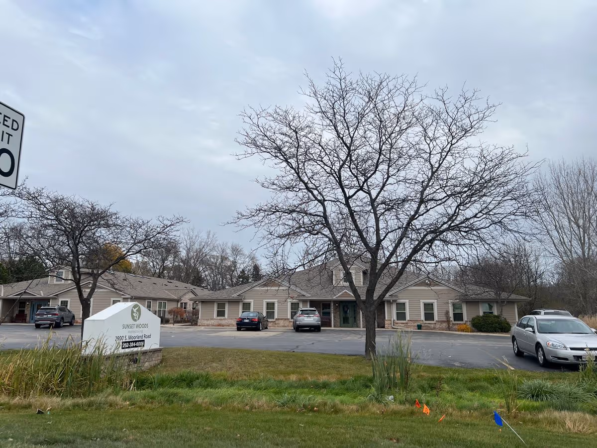 Single-story Sunset Woods senior living building with parked cars, leafless trees, and a front sign under an overcast sky.