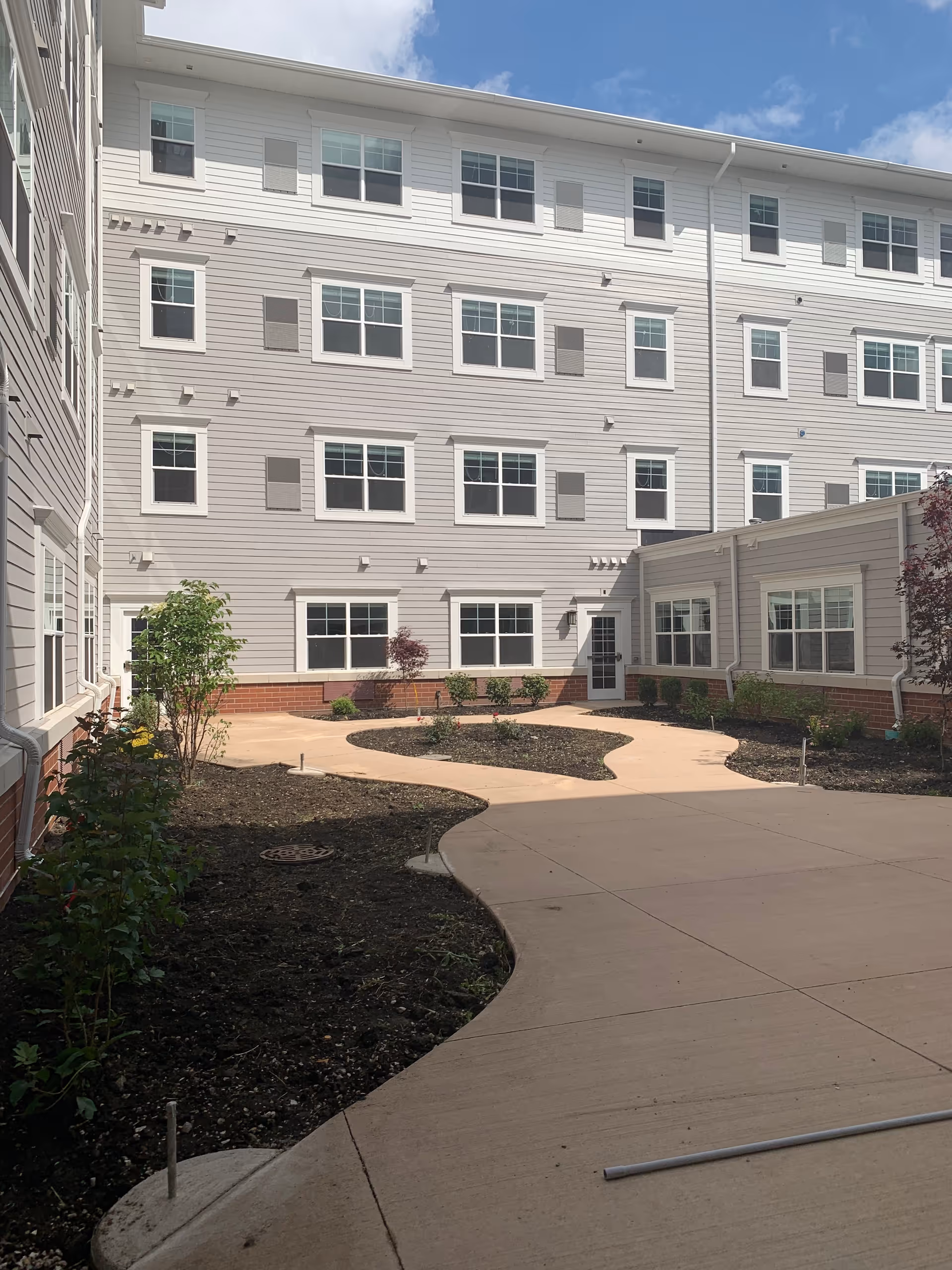 Outdoor courtyard area of a senior living facility with a winding concrete pathway, newly planted shrubs and small trees, surrounded by a multi-story building with many windows under a partly cloudy sky.