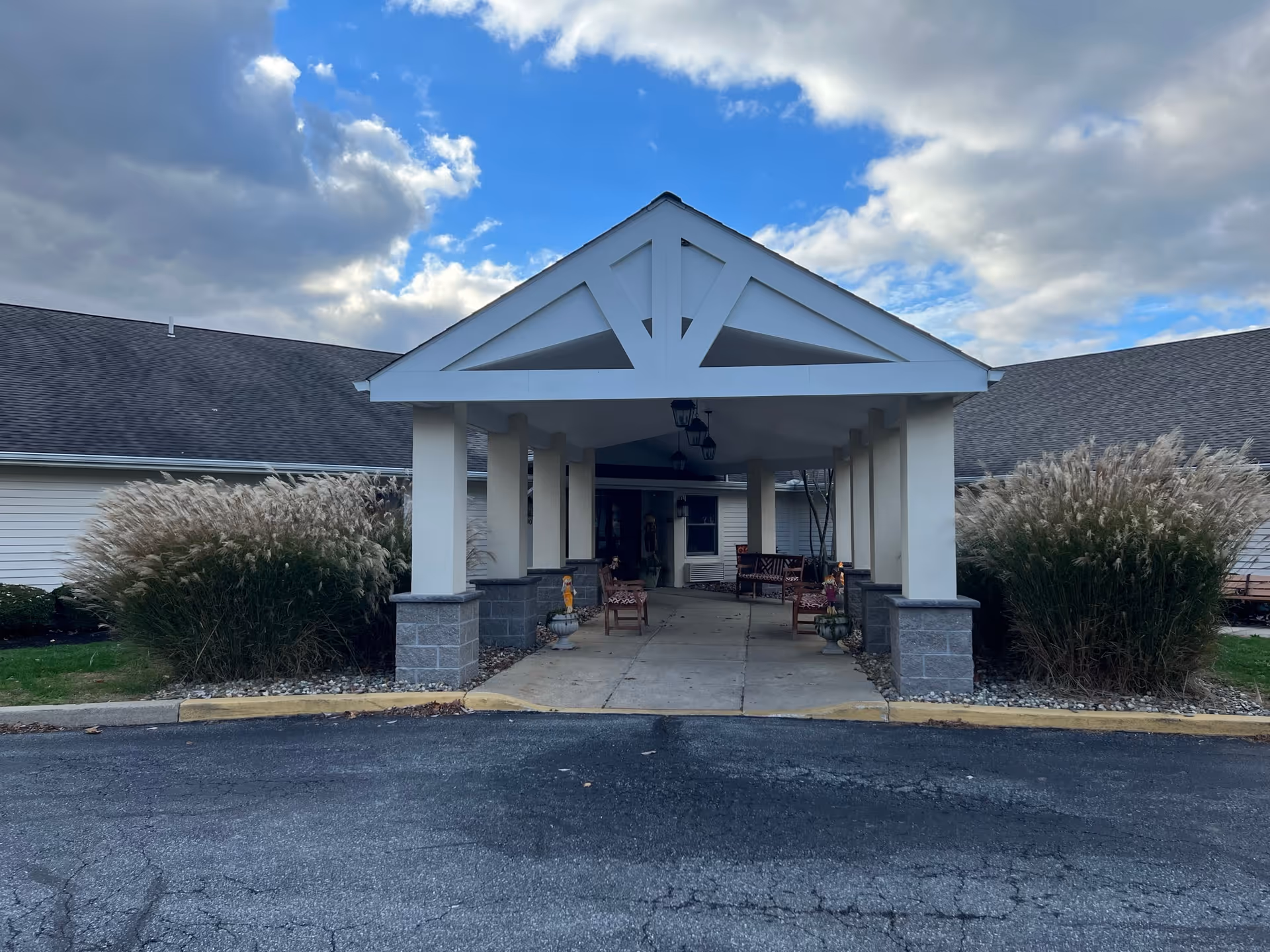 Covered entrance (porte-cochère) of a single-story senior living facility with benches and ornamental grasses under a partly cloudy sky.
