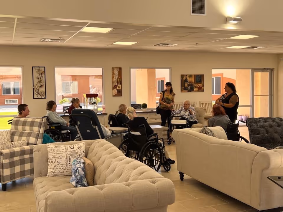 A group of elderly individuals, some in wheelchairs, gathered in a spacious, well-lit assisted living facility common area with beige sofas and armchairs. Two women are standing and engaging with the seated residents. Large windows and glass doors allow natural light to fill the room, and decorative wall art is visible.