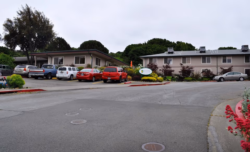 Exterior view of Greenridge Senior Living facility showing two beige buildings with multiple windows, a parking area with several cars, and landscaped greenery including bushes and trees under an overcast sky.