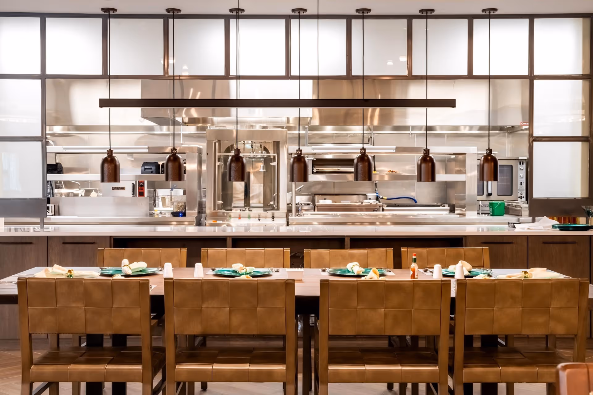 A modern dining area with a long wooden table set with green plates, napkins, and silverware. Brown leather chairs surround the table. Behind the table is a large open kitchen with stainless steel appliances and equipment, visible through glass panels. Several pendant lights hang above the table.