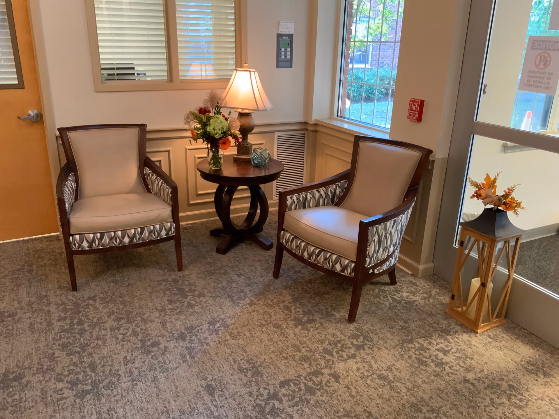Two upholstered chairs flanking a round table with a lamp and floral arrangement in a small lobby seating area by a glass door.