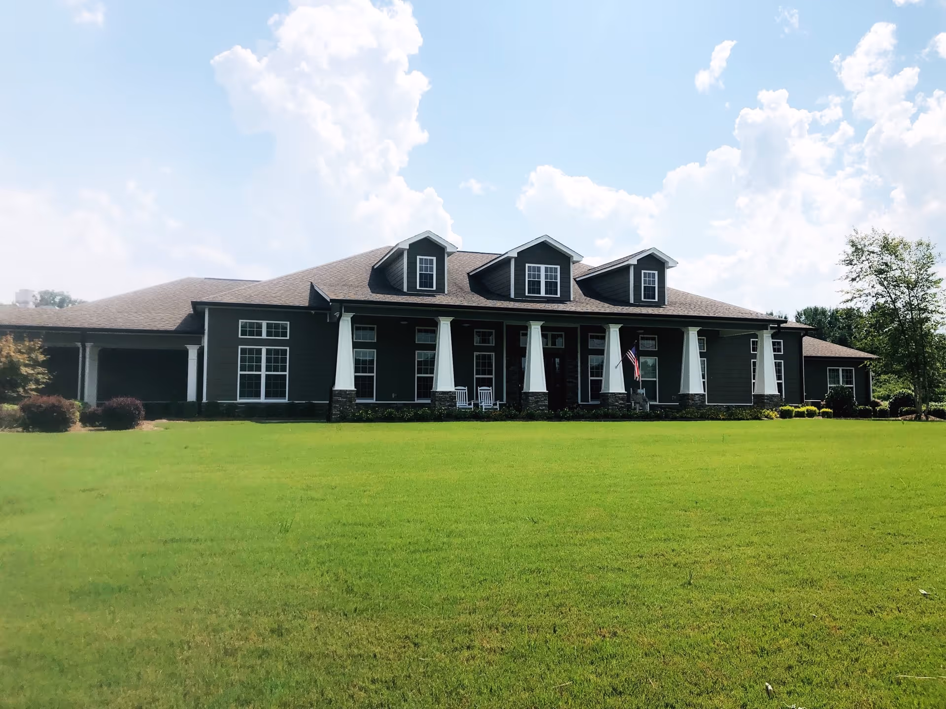 Front exterior of a single-story residential building with a wide porch, columns, dormer windows, and a large green lawn.