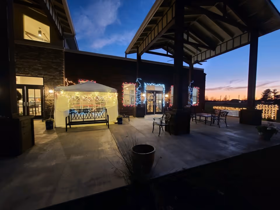Covered entrance patio of Webwood Assisted Living at dusk with string lights, a lit canopy, benches and tables near glass doors.