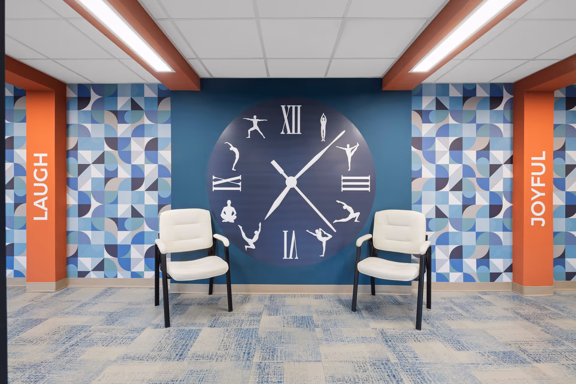 Two white chairs sit in front of a large clock mural on a blue wall flanked by geometric patterned wallpaper and orange columns labeled 'LAUGH' and 'JOYFUL'.