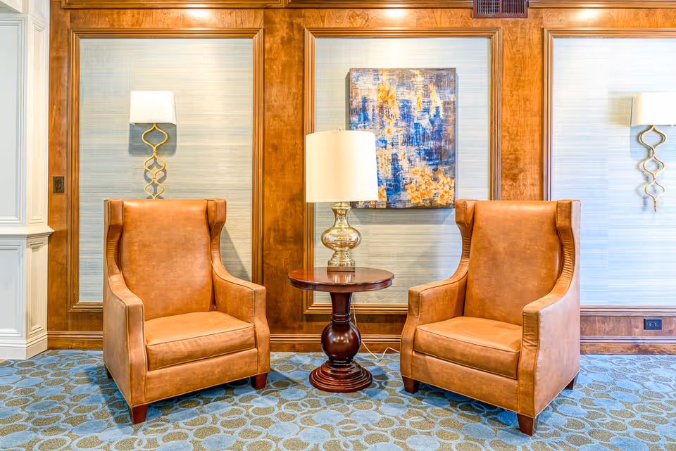 Two brown leather armchairs with high backs are positioned on either side of a round wooden side table with a silver lamp on it. The background features wood paneling with light blue textured wallpaper and a colorful abstract painting in the center. Two wall sconces with white shades and gold bases flank the chairs on the wall.