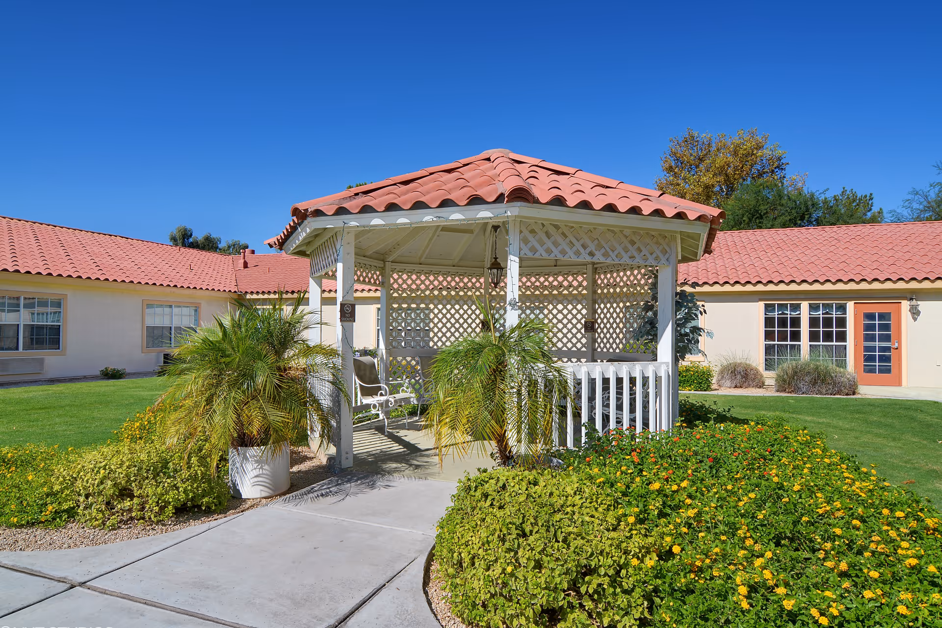 A white gazebo with a red tiled roof situated in a garden area with green grass, bushes, and flowering plants. The gazebo has lattice walls and a hanging lantern inside. Surrounding the gazebo are buildings with red tiled roofs and beige walls under a clear blue sky.