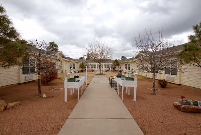 Outdoor courtyard area at Avista Senior Living Payson featuring a concrete walkway flanked by raised garden beds and benches, with single-story beige buildings on either side and leafless trees under a cloudy sky.
