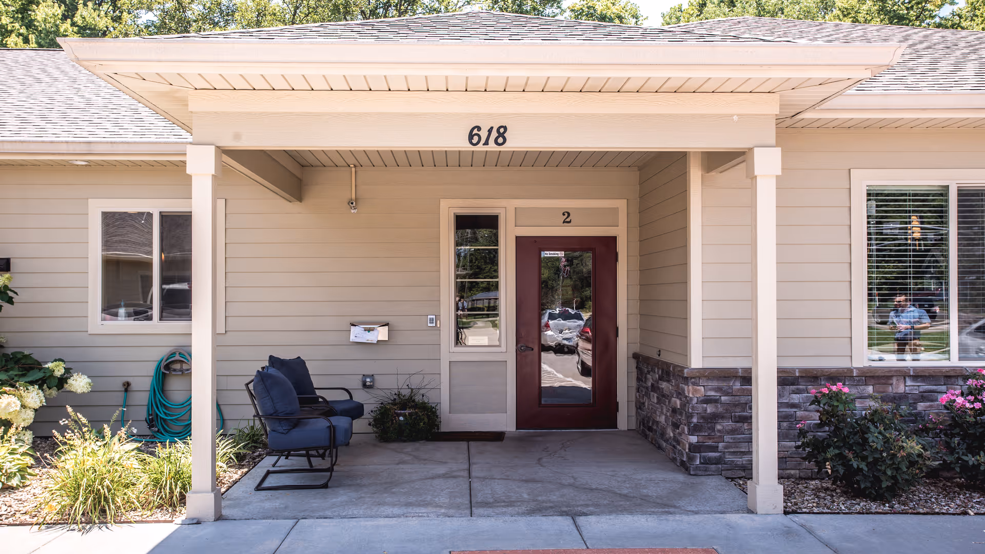 Front covered entrance of a single-story senior living building with the number 618 above a maroon door, a pair of chairs, and landscaping.