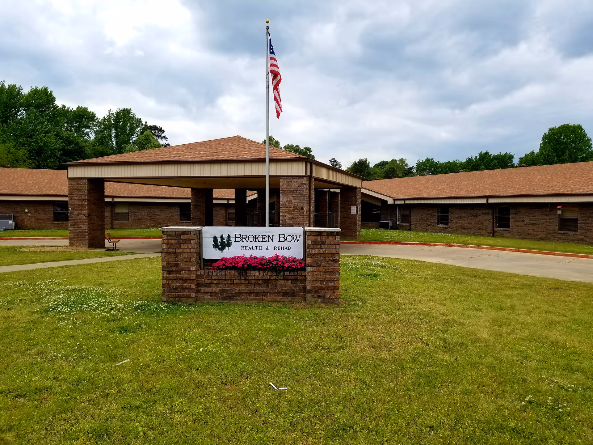 Front exterior view of Broken Bow Health and Rehab building with a brick entrance structure, an American flag on a pole, and a sign displaying the facility name surrounded by green grass and trees under a cloudy sky.