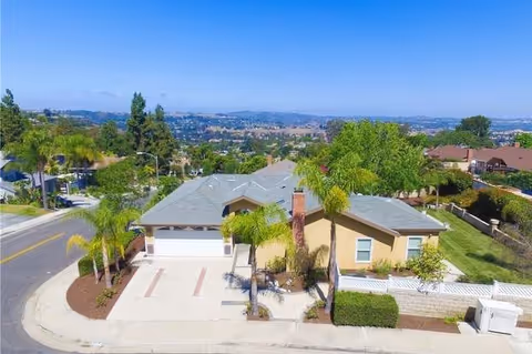 Aerial view of a single-story residential building with a gray roof and beige exterior, surrounded by palm trees and greenery, located on a corner lot with a driveway and a street visible.