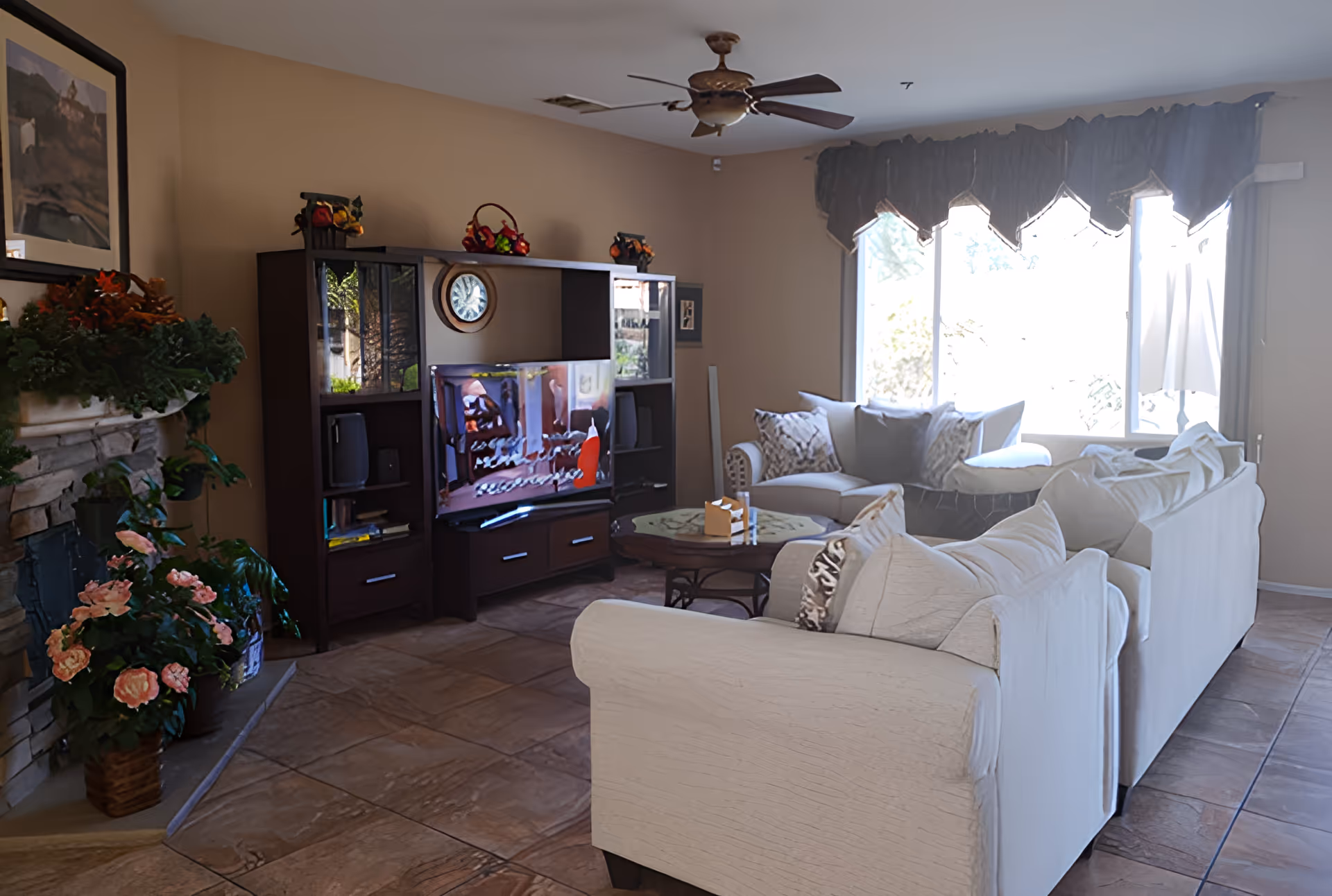 A cozy living room with beige walls and tiled floor featuring two white sofas with decorative pillows, a round coffee table, a dark wood entertainment center with a TV, a ceiling fan, a large window with dark valances, and a stone fireplace adorned with plants and flowers.