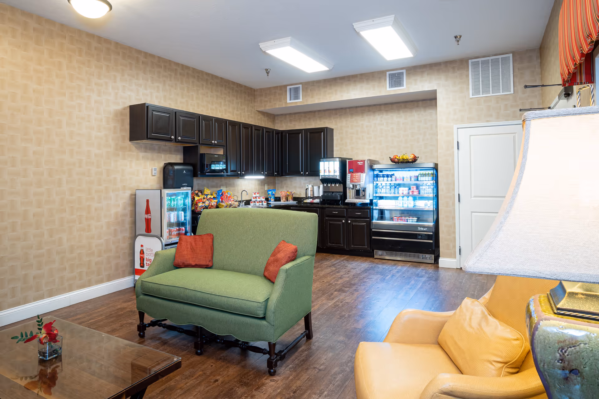 Interior view of a common area with a small kitchenette featuring dark cabinets, a mini fridge, a beverage dispenser, and a refrigerated display case. In the foreground, there is a green loveseat with two red pillows, a yellow armchair with a matching pillow, a table lamp, and a wooden coffee table with a small flower arrangement.