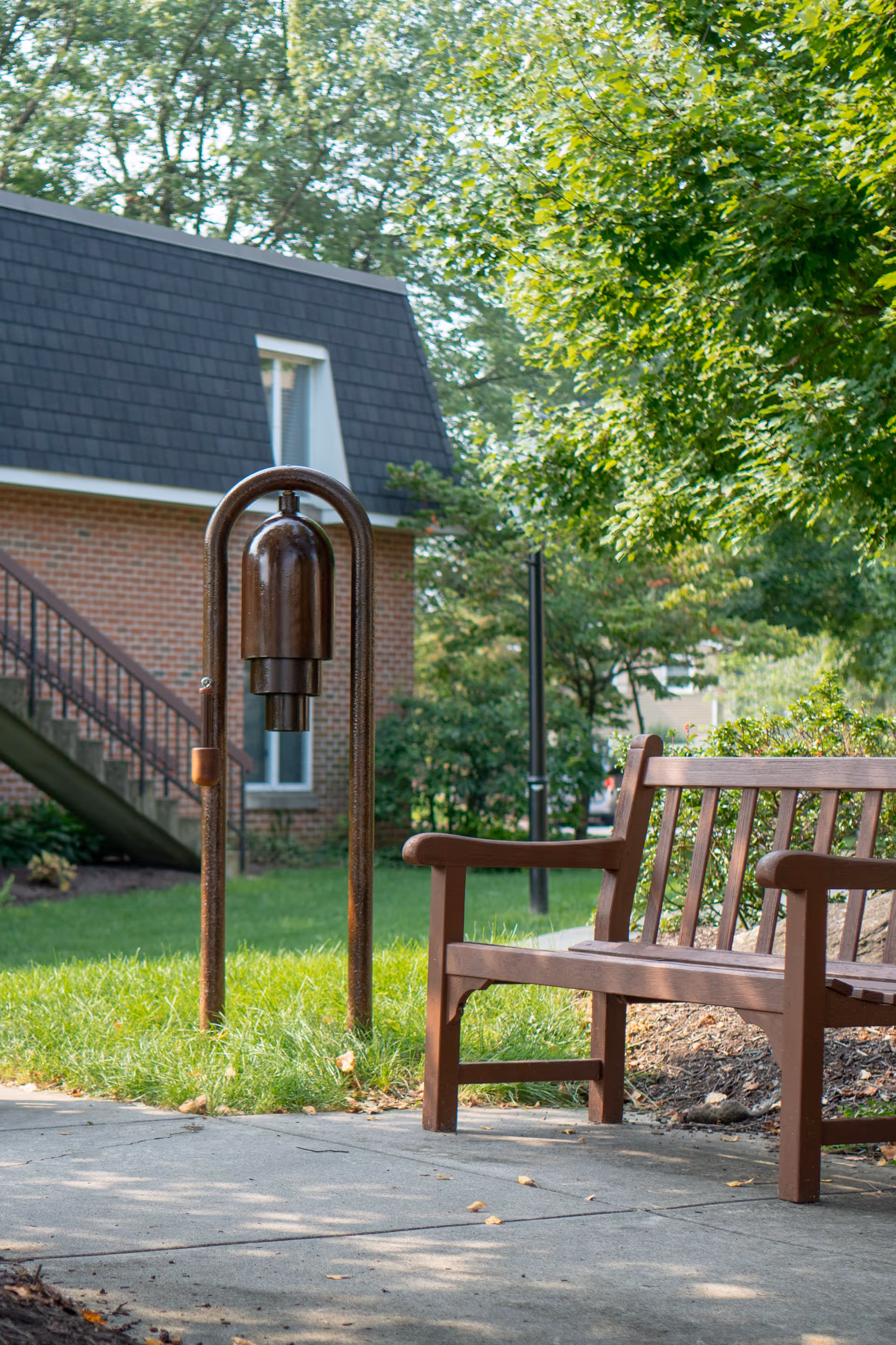 Outdoor scene at Moravian Manor Communities showing a brown wooden bench on a paved walkway next to a large metal bell mounted on a frame. In the background, there is a brick building with a staircase and green trees providing shade.