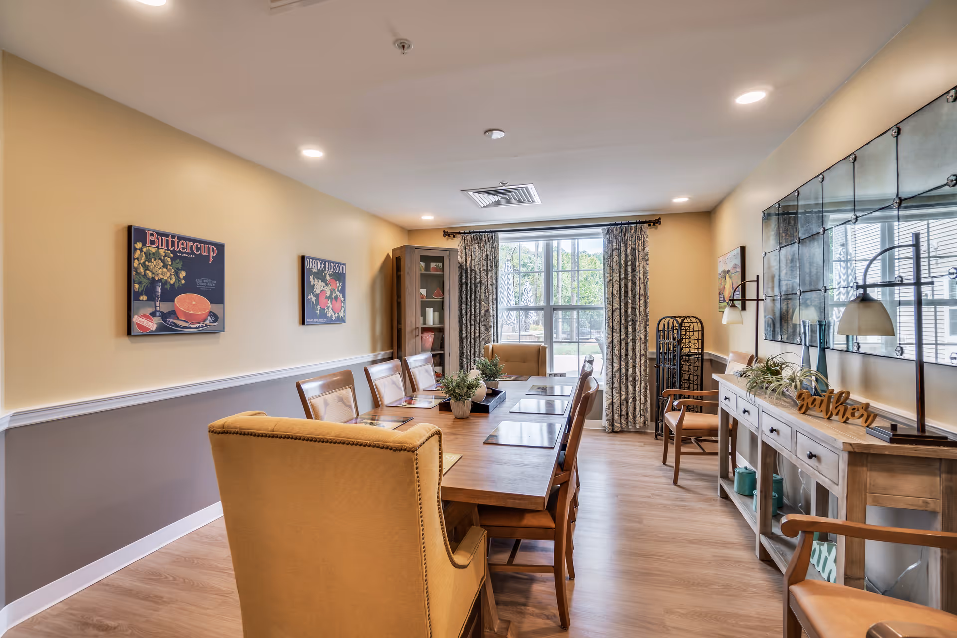 A well-lit dining room with a long wooden table surrounded by chairs, including two upholstered armchairs at the ends. The walls are painted in a two-tone style with yellow on top and gray on the bottom, decorated with framed floral artwork. A large window with patterned curtains lets in natural light. On the right side, there is a wooden sideboard with decorative items, including a sign that says 'gather' and two table lamps. The floor is light wood, and the room has a cozy, inviting atmosphere.
