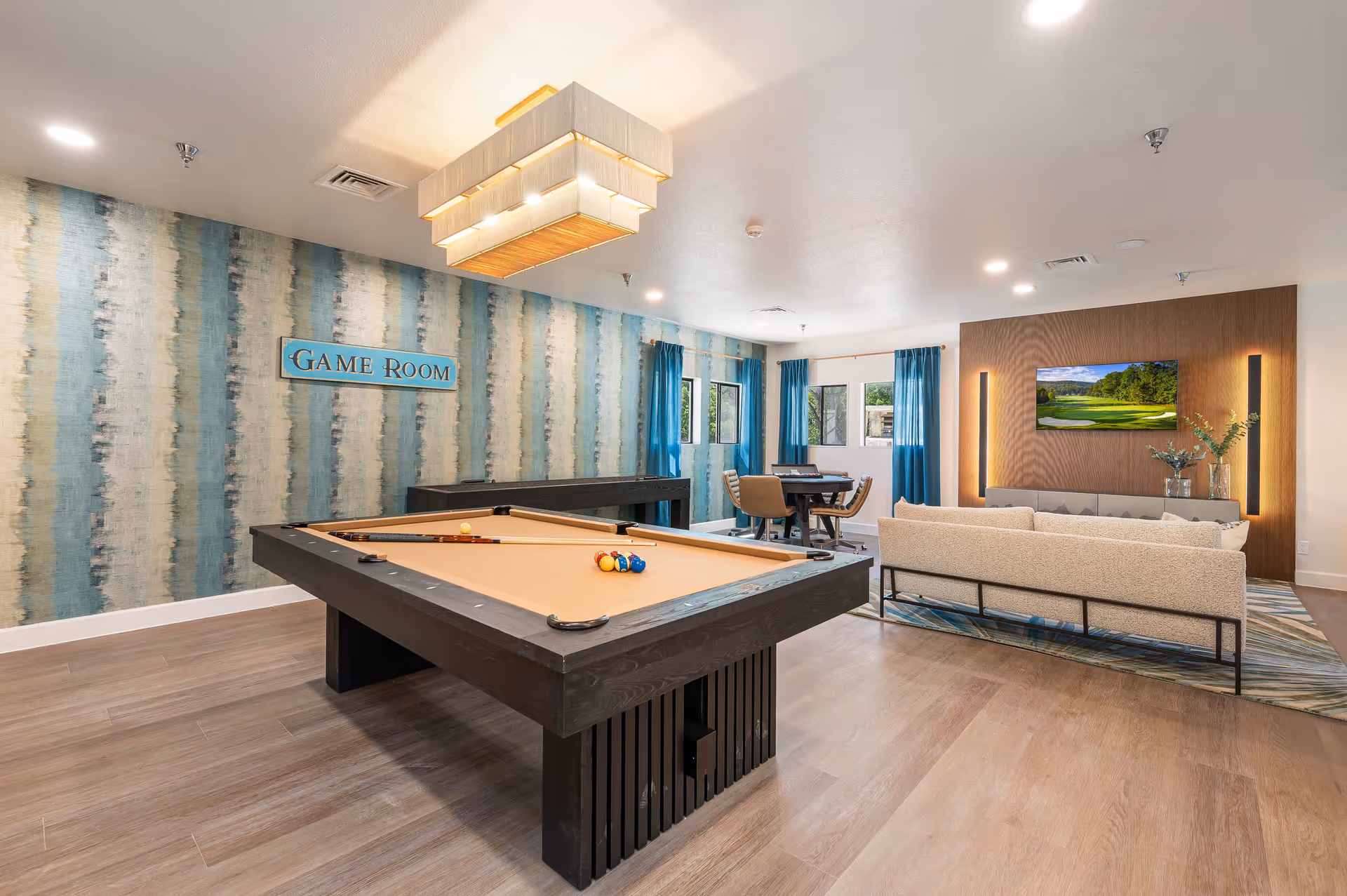 A bright and spacious game room featuring a pool table with cues and balls, a shuffleboard table against a blue and white striped wall with a 'Game Room' sign, a seating area with a beige sofa facing a wall-mounted TV, and a card table with chairs near windows with blue curtains.