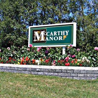 A green and white sign for McCarthy Manor surrounded by colorful flowers and greenery, with trees in the background.