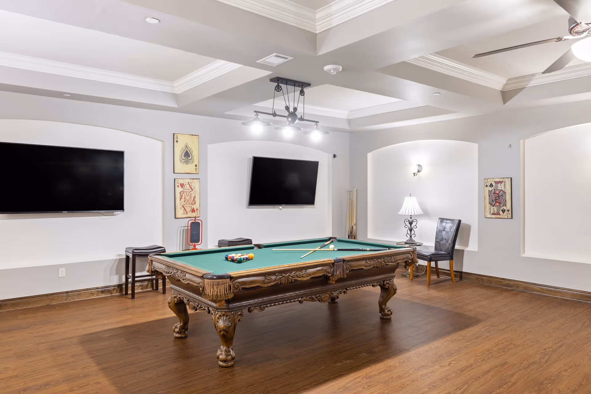 A well-lit game room with a classic wooden pool table in the center, two wall-mounted flat-screen TVs, decorative playing card artwork on the walls, a leather chair, and a table lamp in a recessed wall niche.